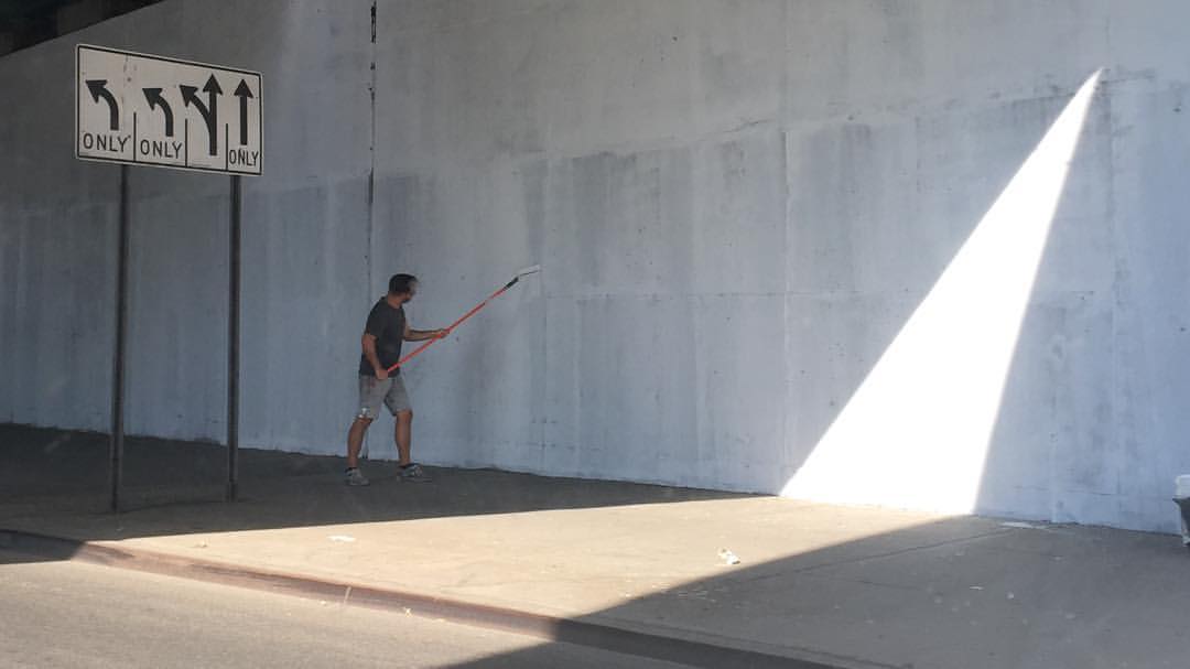 Adult male painting a concrete wall near a street, using a paint roller, with a road sign in the background.