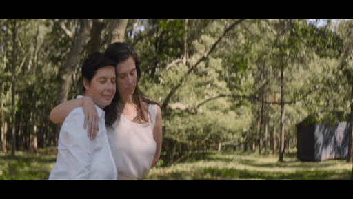 Two adult females embrace in a sun-dappled outdoor setting with a wooded area and a dark shed in the background, creating a serene moment.