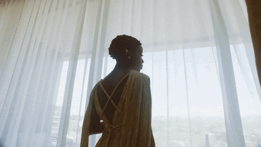 Woman in a gown looks out a window with sheer white curtains. Her back is to the viewer, and a city is visible through the window.