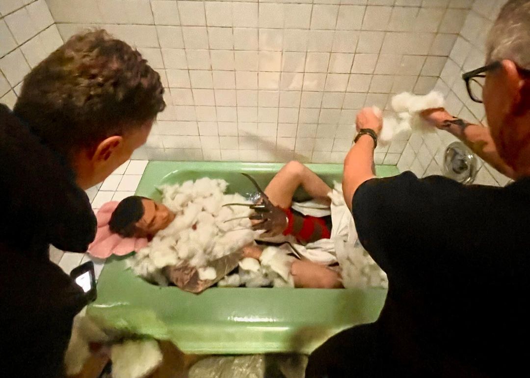 Man in bathtub covered with cotton, styled as Freddy Krueger, with two men looking on in a bathroom with green tub and white tile.
