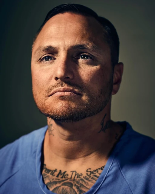 Portrait of a man with short dark hair, stubble, and visible tattoos on his neck and chest. He is wearing a blue shirt.