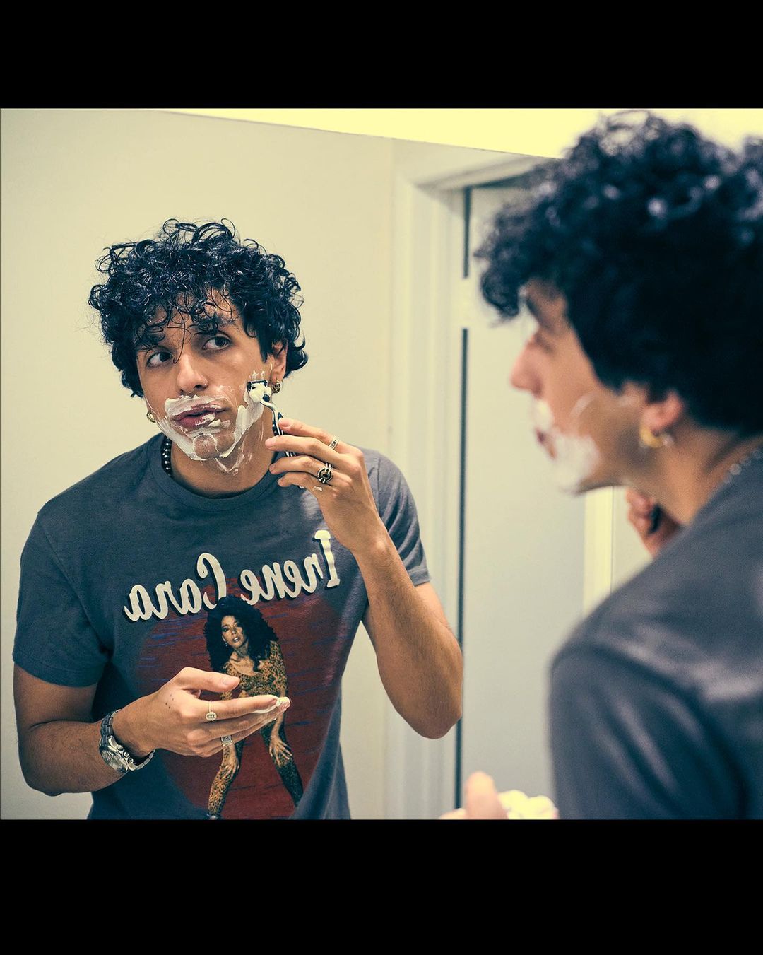 Adult male shaves with razor in mirror. Dark curly hair, t-shirt with female portrait. Bathroom setting. Rings and beaded necklace visible.