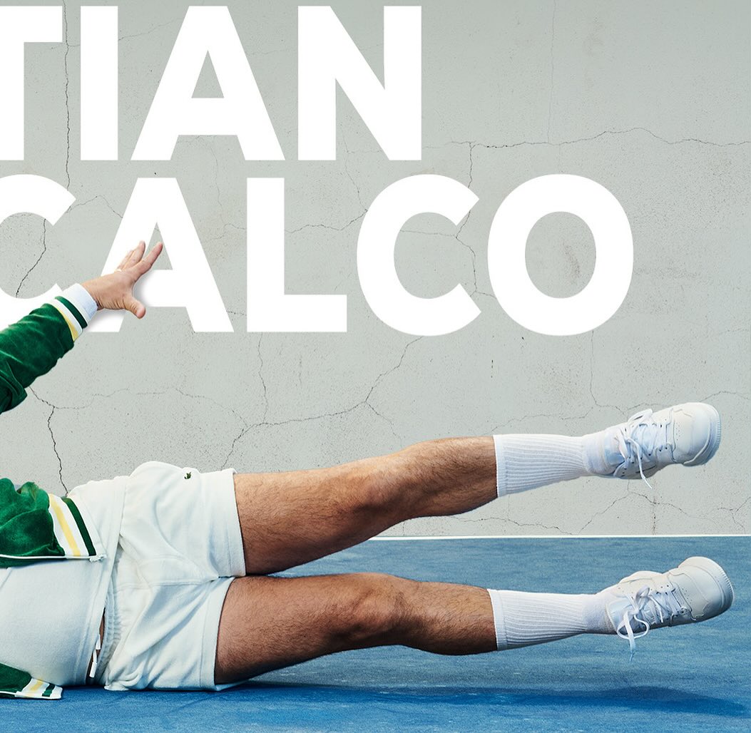 Athletic male poses dynamically on a blue surface with white text overlay, wearing vintage sportswear, white shorts, socks, and sneakers.