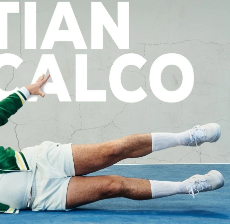 Athletic male poses dynamically on a blue surface with white text overlay, wearing vintage sportswear, white shorts, socks, and sneakers.