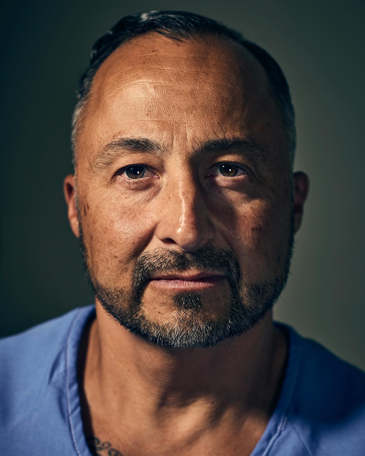 Close-up portrait of a middle-aged Hispanic man with short hair, a trimmed beard, and a tattoo on his chest wearing a blue collared shirt.