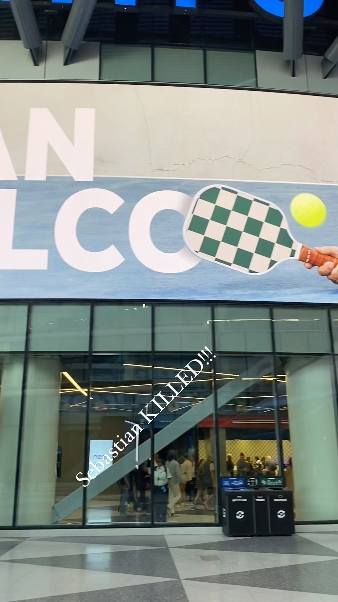 Indoor shot featuring a convention hall entrance with a large banner displaying a pickleball paddle graphic, captured in bright daylight.