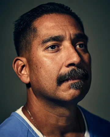 Close-up portrait of a man in his 40s with a dark mustache, wearing a blue shirt against a dark background