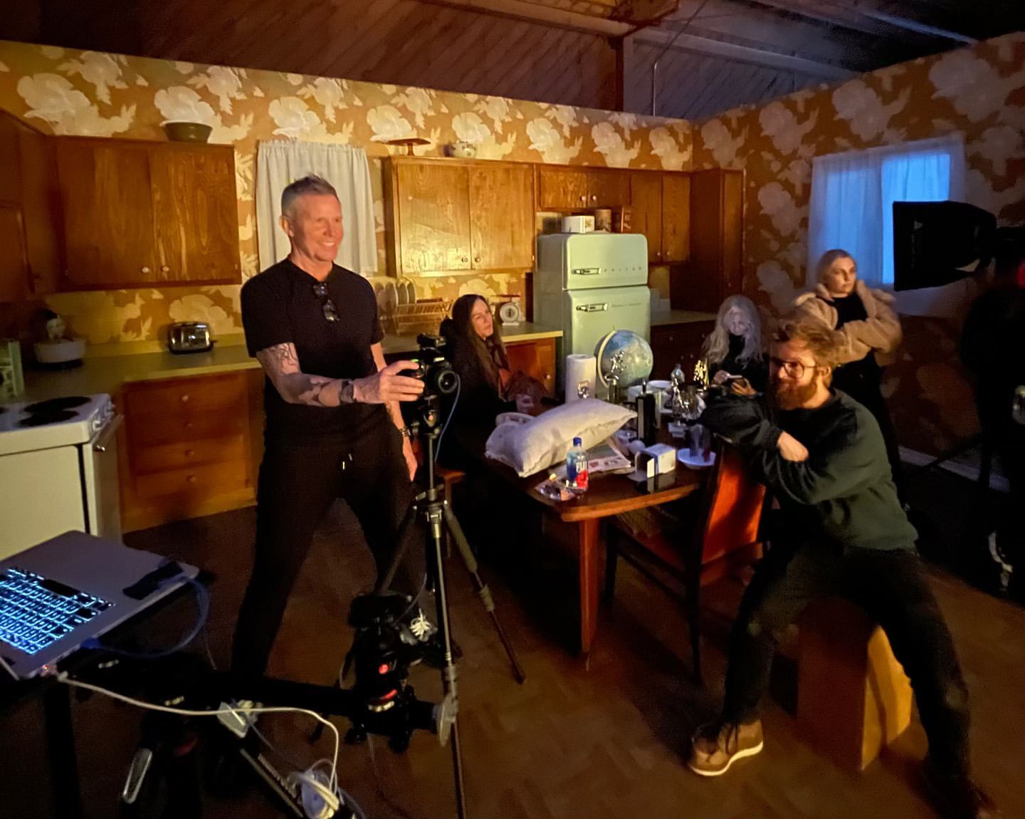On a video set, a male videographer stands near a camera on a tripod, while other people sit around a table in a vintage-style kitchen.