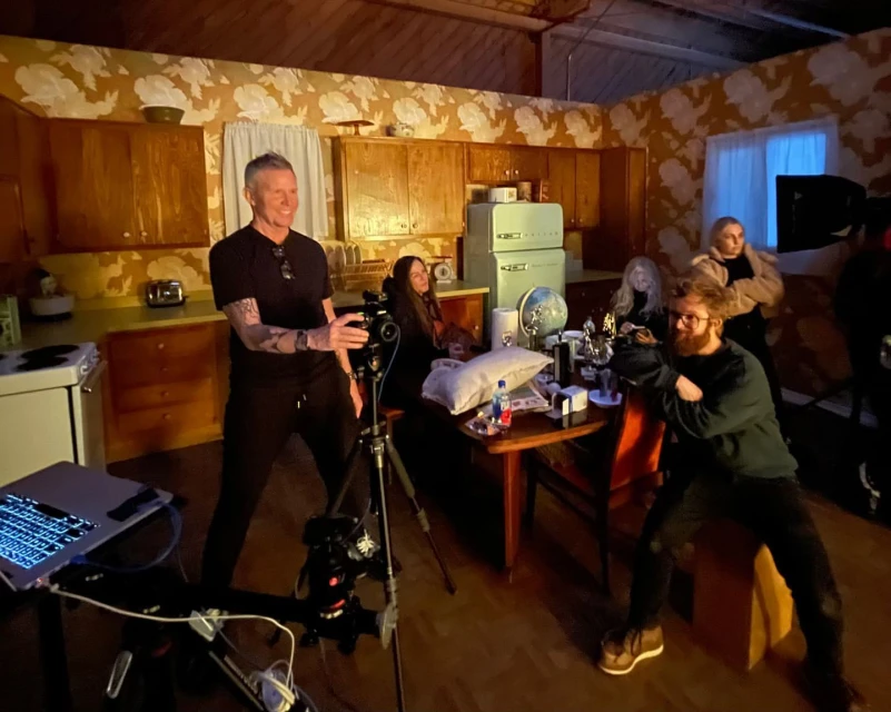 On a video set, a male videographer stands near a camera on a tripod, while other people sit around a table in a vintage-style kitchen.