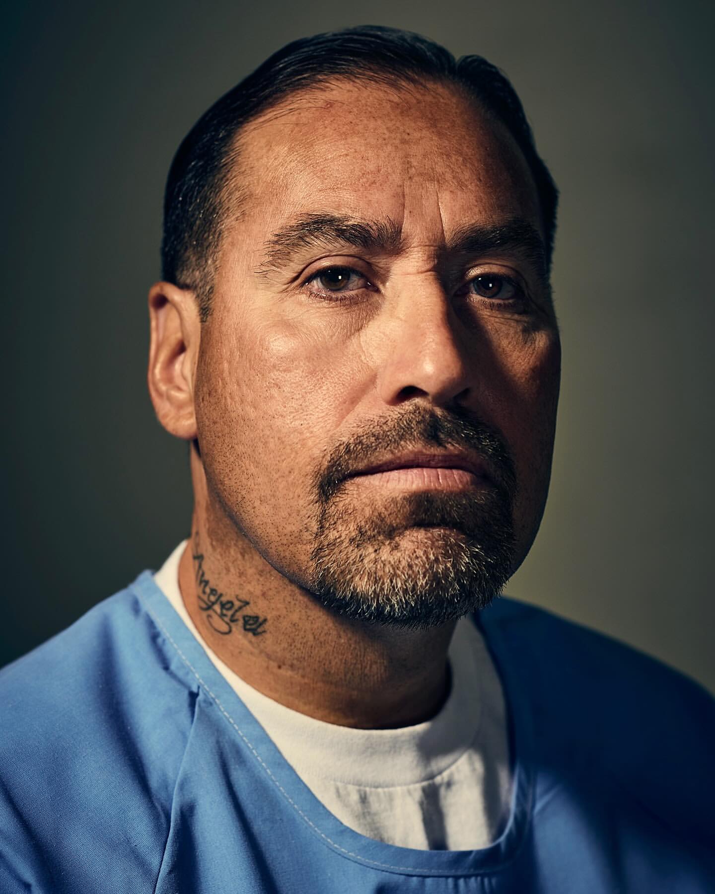 Close-up portrait of a man with a goatee wearing a blue scrub top, facing forward with a serious expression, and a gray background.