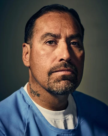 Close-up portrait of a man with a goatee wearing a blue scrub top, facing forward with a serious expression, and a gray background.