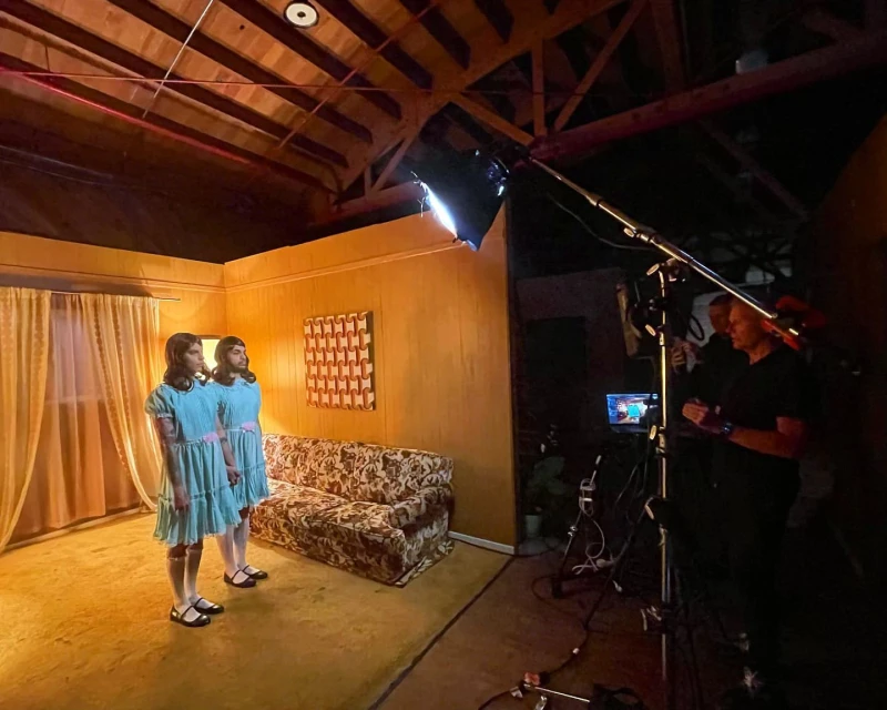 Two young women in matching blue dresses and black shoes stand in a retro-style room with floral couch, captured during a film shoot.