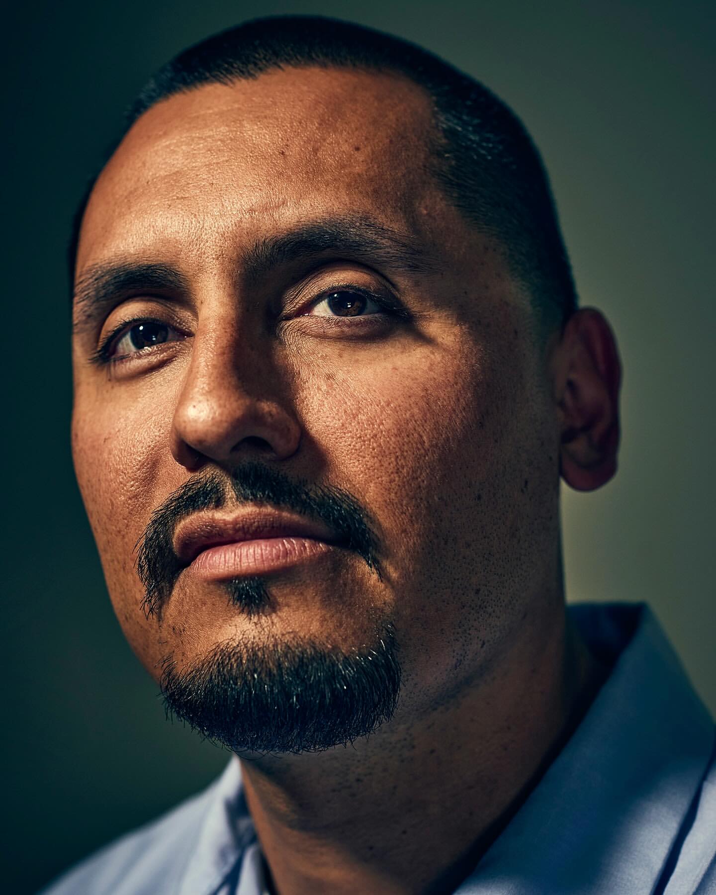 Close-up portrait of a middle-aged Hispanic man with a goatee wearing a blue button-down shirt against a dark, neutral background.
