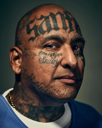 Close-up portrait of adult male with face and neck tattoos, wearing a blue shirt, set against a dark grey background in a studio.