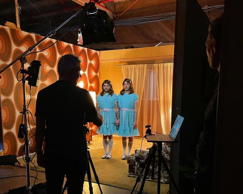 Two actors in matching blue dresses recreate a scene from 'The Shining' on a set with retro wallpaper and studio lighting. Cinematic shot.
