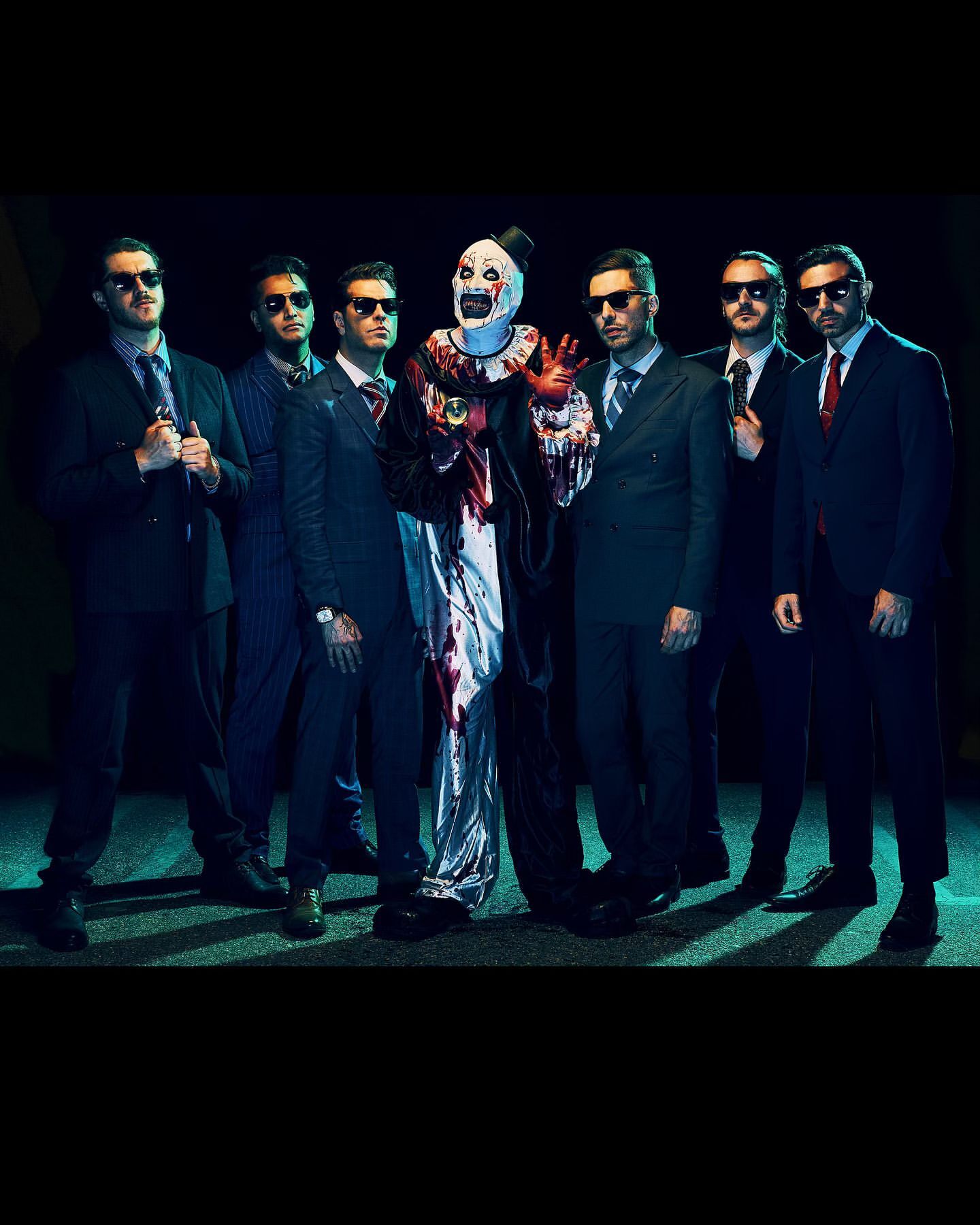 Group of men and a clown in formal attire, standing together in a darkly lit indoor setting, possibly for an event or performance.