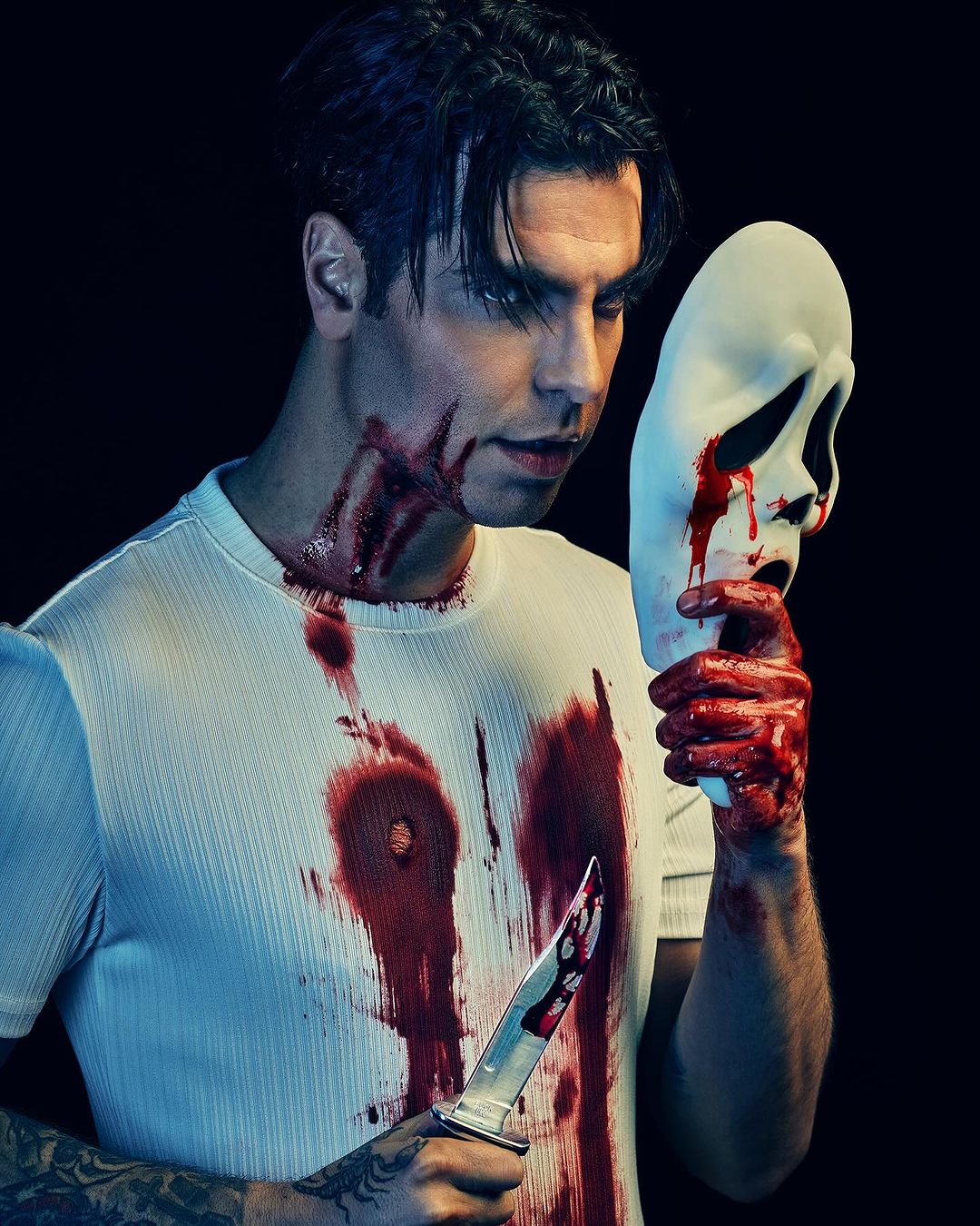 A man in a white shirt holding a bloody knife and a 'Scream' mask, partially covered in blood, against a dark background.