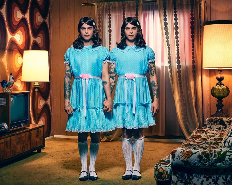 Two adults dressed as twins in matching blue dresses and white socks, standing in a retro-styled living room with patterned wallpaper.