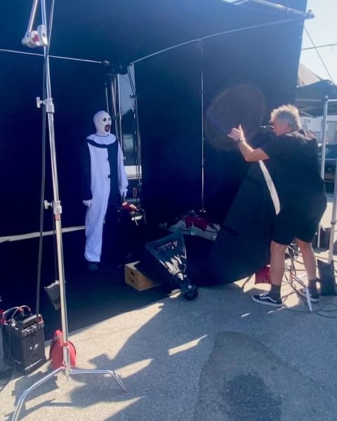 Behind-the-scenes shot of a performer in black and white clown costume and mask being filmed on a sound stage, with lighting and production crew.