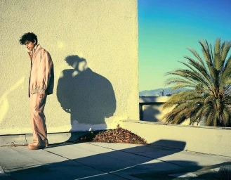 Man with curly hair wearing a tan jacket and trousers standing on a rooftop with his shadow cast against a textured yellow wall.