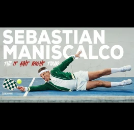 Man wearing a green track jacket and white shorts diving horizontally on a blue tennis court while playing with a green and white paddle.