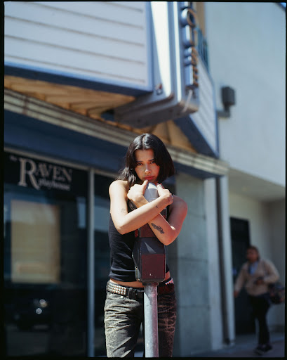 A woman with dark hair stands in front of a building, leaning against a parking meter. The scene takes place outside on a sunny day.