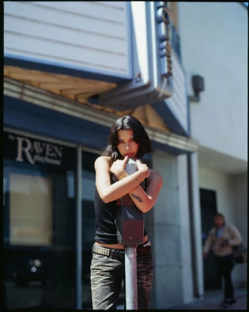 A woman with dark hair stands in front of a building, leaning against a parking meter. The scene takes place outside on a sunny day.