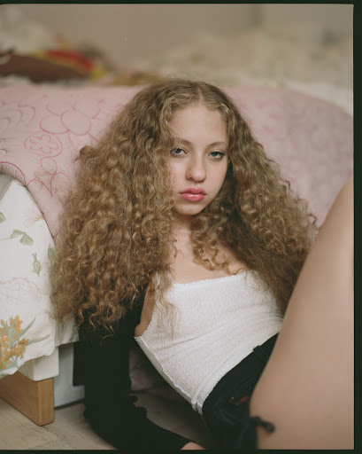 Young woman with curly blond hair, wearing a white top and black jacket, posing indoors with a slight frown, looking at the camera.