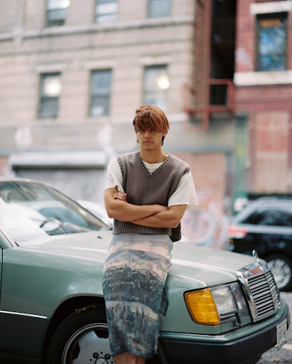 Adult male posing outdoors wearing a sweater vest and landscape-printed skirt, leaning on a light blue Mercedes-Benz in the city.