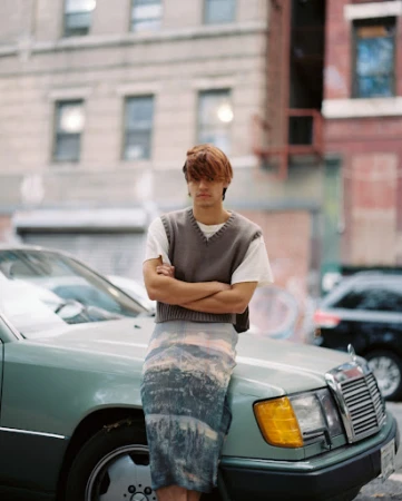Adult male posing outdoors wearing a sweater vest and landscape-printed skirt, leaning on a light blue Mercedes-Benz in the city.