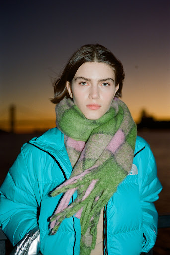 Young woman poses outdoors in a bright blue puffer jacket and green and pink plaid scarf against a twilight sky with blurred background.