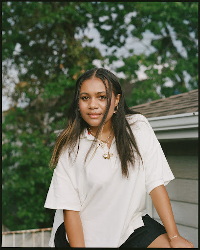 Portrait of an adult woman with braided hair, wearing a white collared shirt and dark shorts, outside with trees and sky in background.
