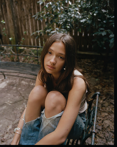 Portrait of a young woman with long brown hair, wearing ripped jeans, sitting on a bench in a backyard with greenery behind her.