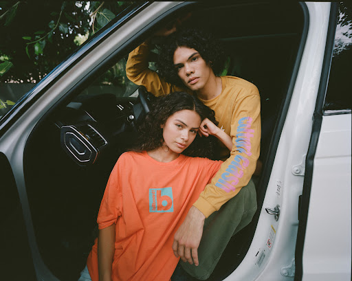 Two young adults, a man and woman, pose in a white car, both wearing casual, branded shirts, creating a relaxed, lifestyle fashion image.