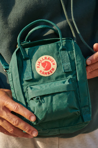 Close-up of a person holding a small, green Fjallraven Kanken backpack purse, with the brand's circular logo visible on the front.