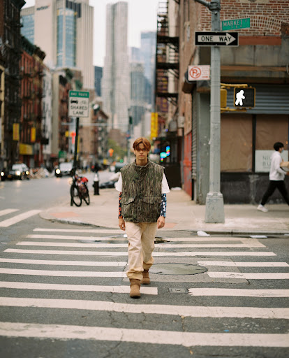Adult male walking on a zebra crossing wearing vest and boots in an urban environment with high-rise buildings in the background.