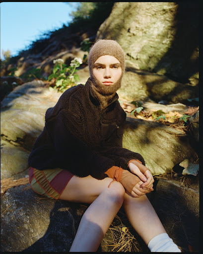 Young woman with knitted balaclava and shorts sits on a rock outdoors, wearing a brown fleece jacket, and white socks surrounded by leaves.