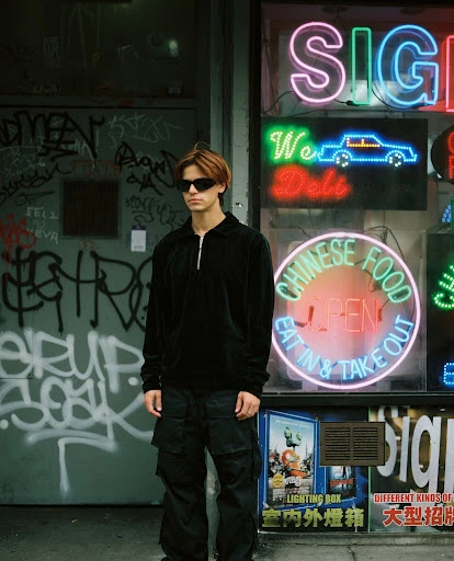 A young adult stands in front of a neon-signed Chinese food restaurant. The man is wearing a dark outfit and sunglasses, posing casually.