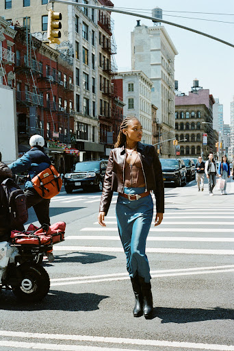 A woman crosses a New York City street in a brown jacket, blue skirt, and black boots on a sunny day with city buildings in the background.