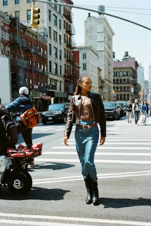 A woman crosses a New York City street in a brown jacket, blue skirt, and black boots on a sunny day with city buildings in the background.