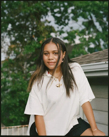 Young woman with long dark braided hair wearing a white polo shirt outdoors by foliage