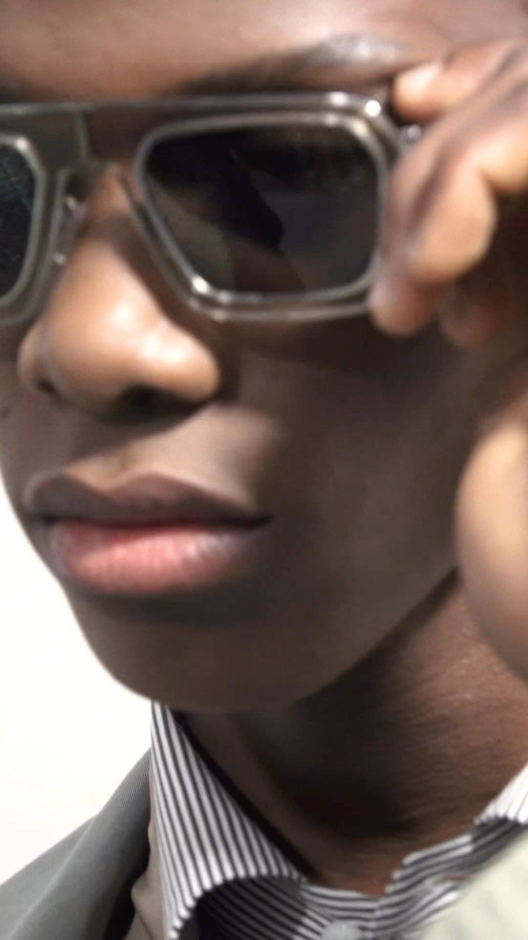 Close-up portrait of an African-American man wearing dark glasses, a gray jacket, and a striped shirt against a plain background.
