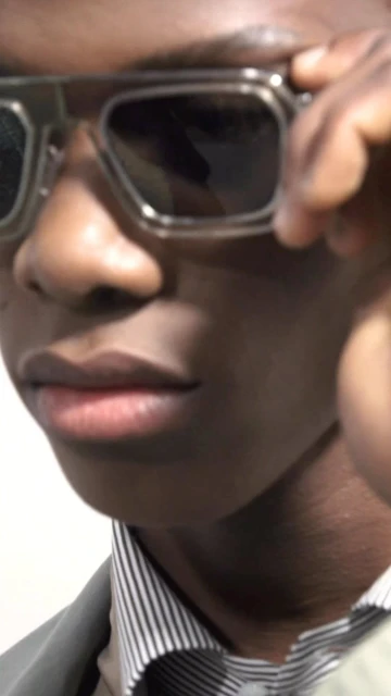 Close-up portrait of an African-American man wearing dark glasses, a gray jacket, and a striped shirt against a plain background.