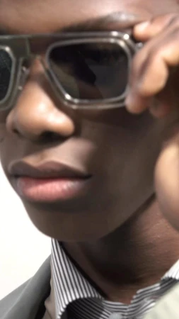 Close-up portrait of an African-American man wearing dark glasses, a gray jacket, and a striped shirt against a plain background.