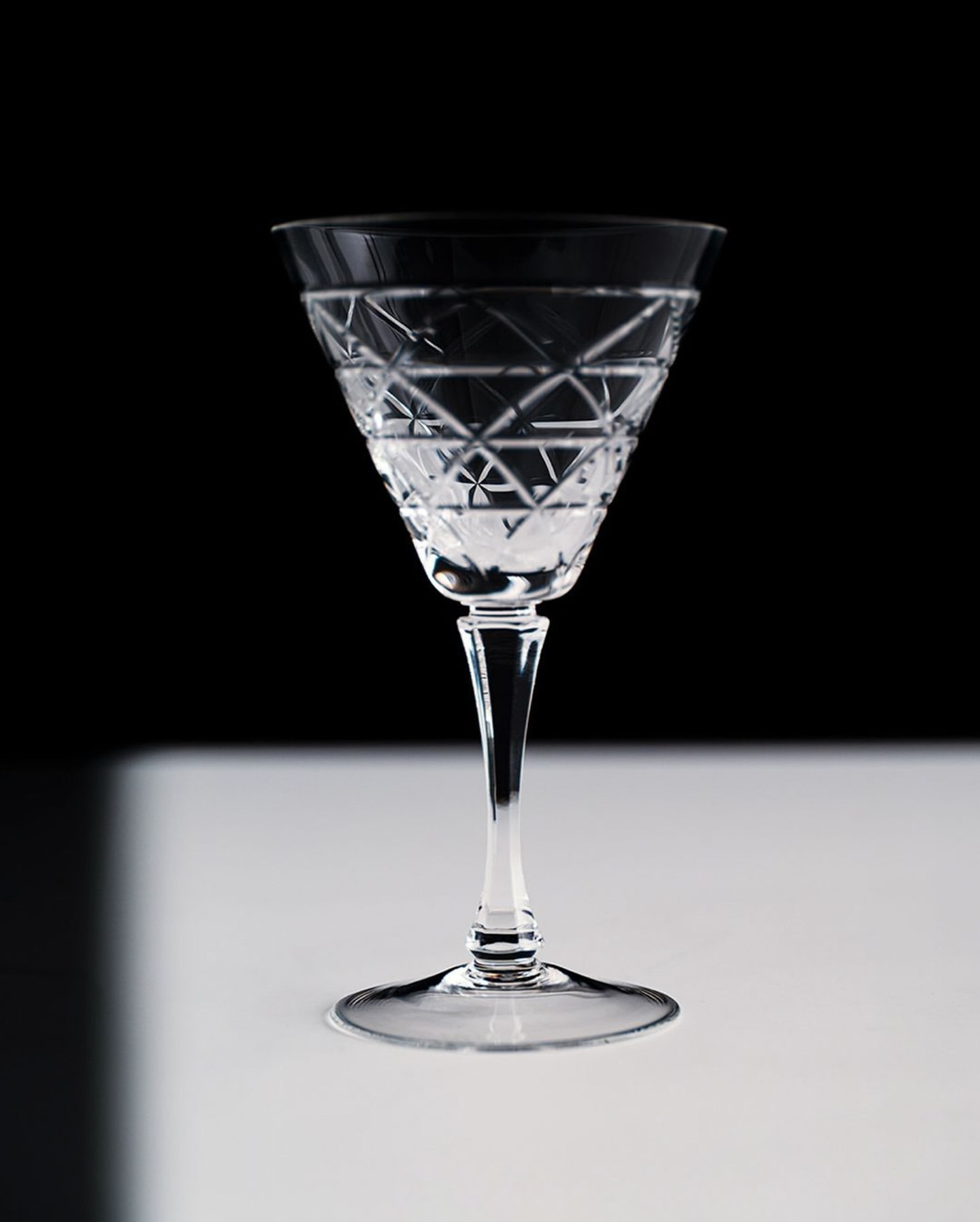Crystal wine glass with intricate diamond pattern stands on a white surface against a stark black background in a studio setting.