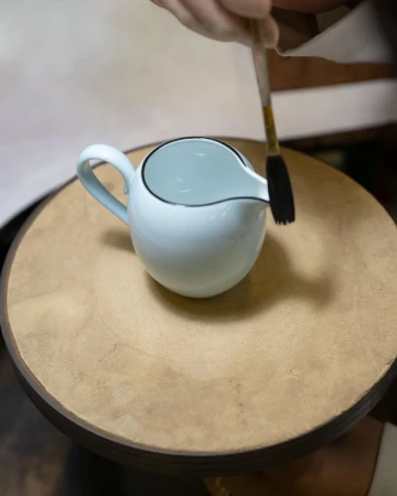 Close-up of a potter painting a white ceramic pitcher on a wooden wheel with a small brush by Michele Foti.