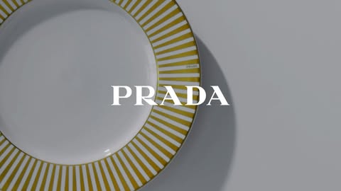 Overhead shot of a white porcelain plate with a gold striped border, the Prada logo is superimposed over the plate on a gray background.