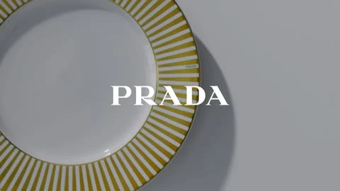 Overhead shot of a white porcelain plate with a gold striped border, the Prada logo is superimposed over the plate on a gray background.