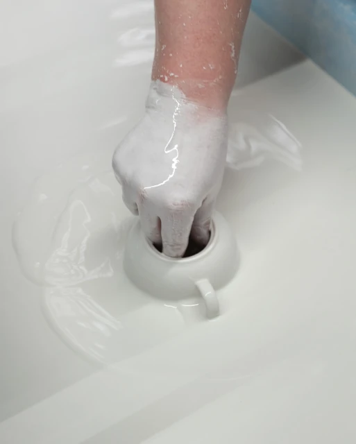 Close-up shot of a hand covered in white slip dipping a ceramic cup into a vat of slip, showcasing the pottery glazing process.