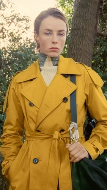 Woman in a yellow trench coat poses outdoors with a green bag, standing near trees, capturing a fashion-forward editorial aesthetic.